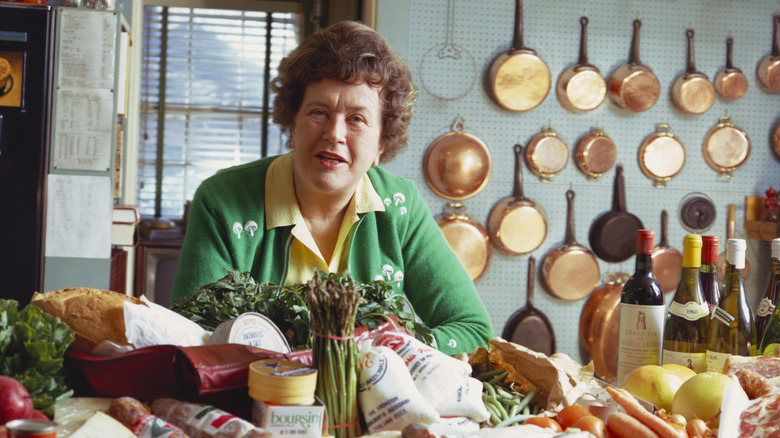 Julia Child in her kitchen with groceries and pots on the wall