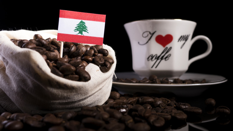 A Lebanese flag is stuck in a bag of coffee beans with a coffee mug behind it.