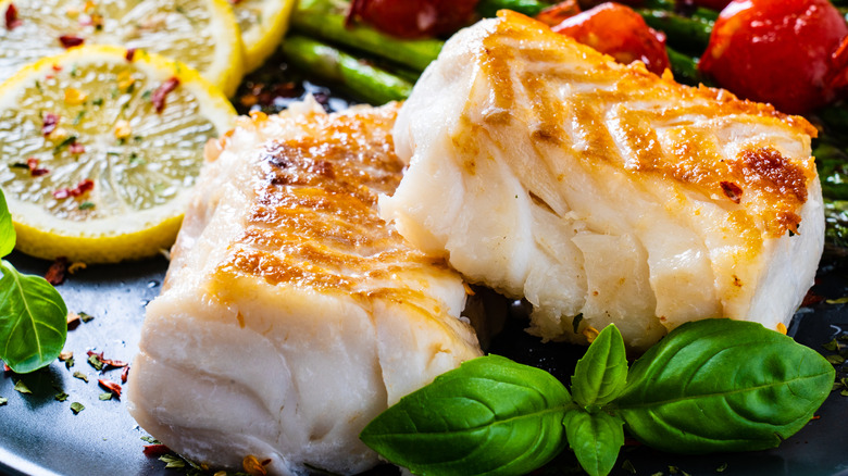 Fresh cooked pieces of roasted cod on a plate of roasted asparagus and tomatoes with lime wheels and fresh basil sprigs
