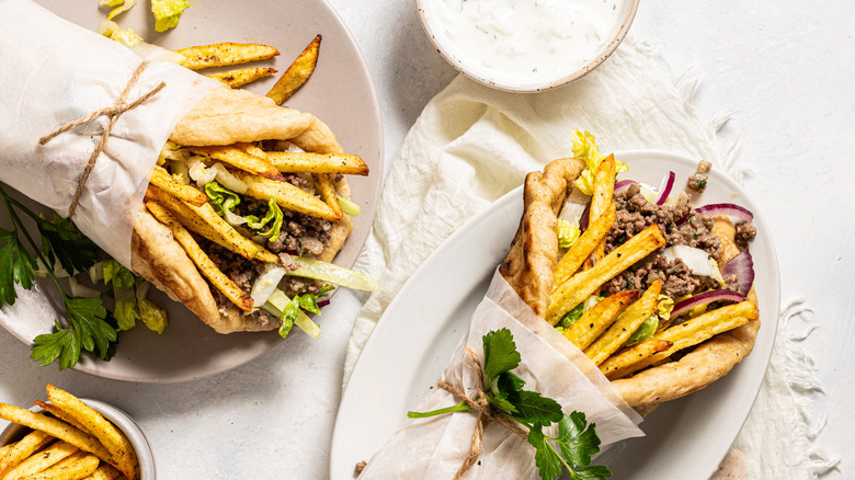 Two wrapped ground beef gyros on plates with a side of tzatziki and fries