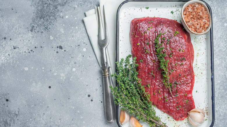 Raw flank steak on a plate with herbs, garlic cloves, and salt