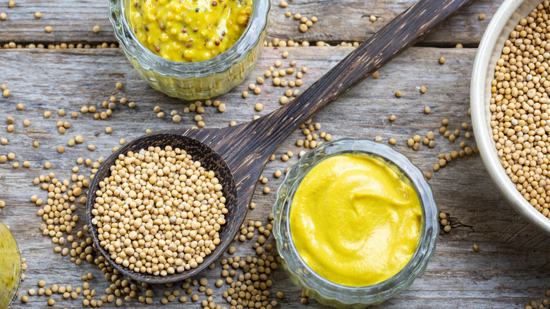 Mustard seeds next to yellow mustard and wholegrain mustard in spoon and bowls, respectively