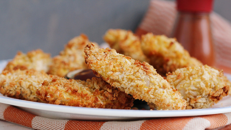 This closeup show panko chicken strips on a plate that rests on a red checkered cloth.