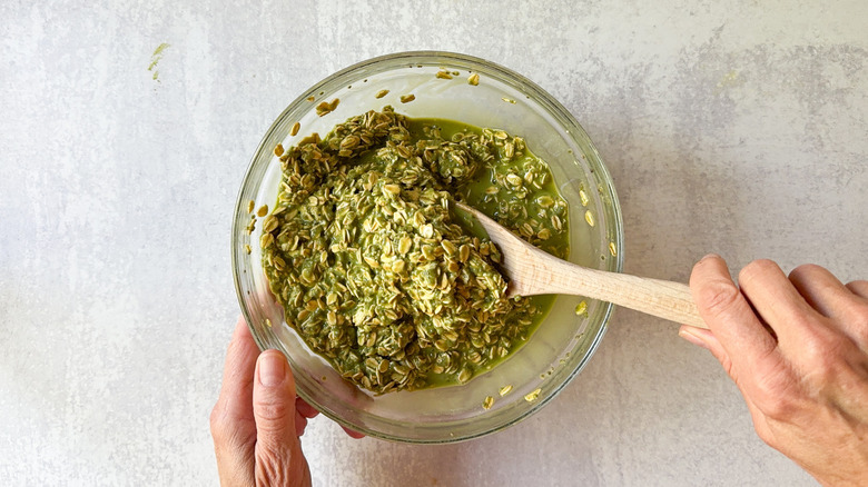 Stirring matcha almond overnight oats in glass bowl with wooden spoon