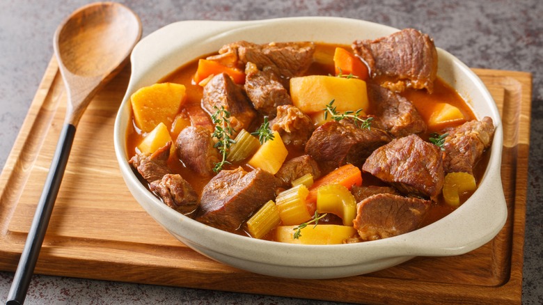 A white bowl of beef stew with meat, potatoes, carrots, celery, and herbs on a wooden cutting board next to a wooden spoon