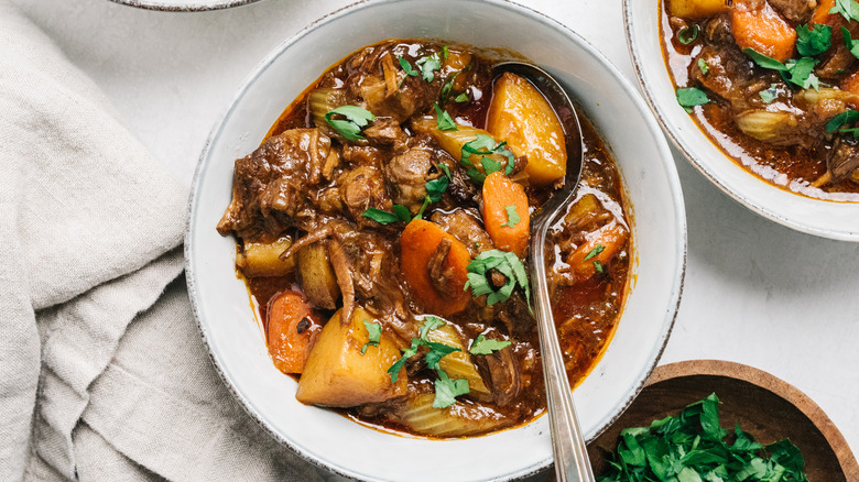 A white bowl of beef stew with meat, potatoes, and carrots with fresh herbs on the top and a silver spoon