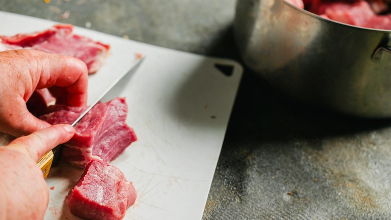 A person cuts raw beef into cubes for beef stew on a white cutting board