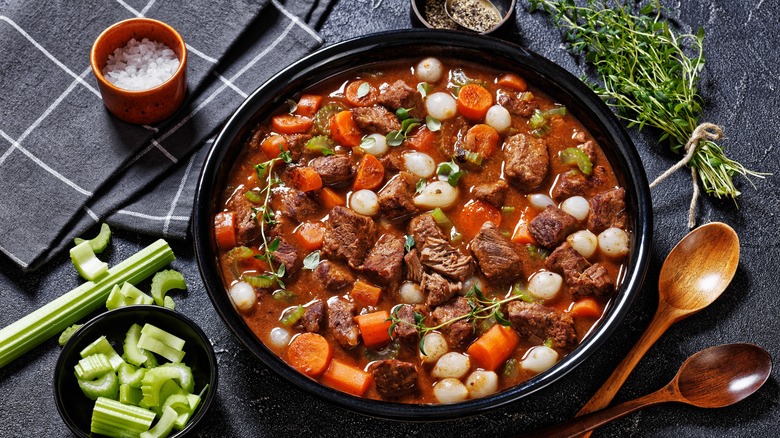 A black bowl of beef stew with pearl onions and carrots with ingredients surrounding the bowl in smaller containers