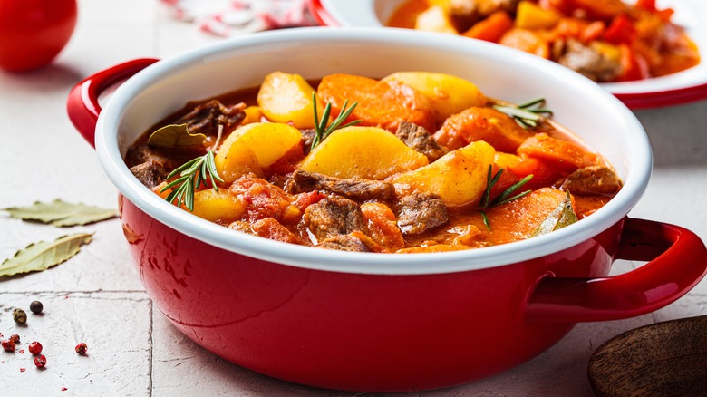 A red bowl of beef stew with large pieces of potatoes and carrots with fresh herbs