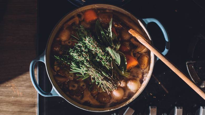 A Dutch oven of beef stew cooking on a stove with fresh herbs on top and a wooden spoon stirring