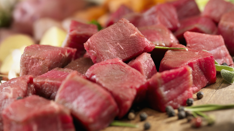 A pile of raw stew meat with herbs and peppercorns on a wooden cutting board