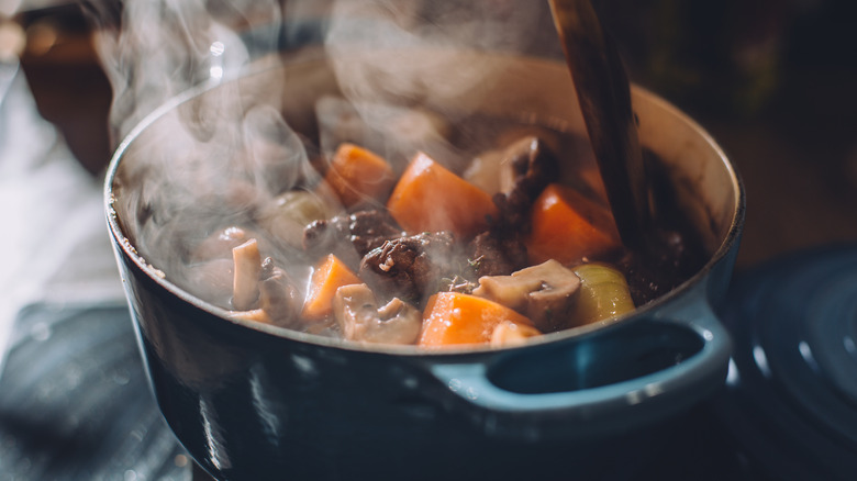 A blue enameled Dutch oven on the stove cooking a pot of beef stew with meat, carrots, and potatoes, stirred by a spoon