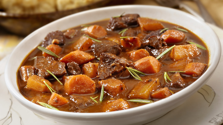 A white bowl full of beef stew with carrots and herbs in rich broth with a white plate with flowers underneath