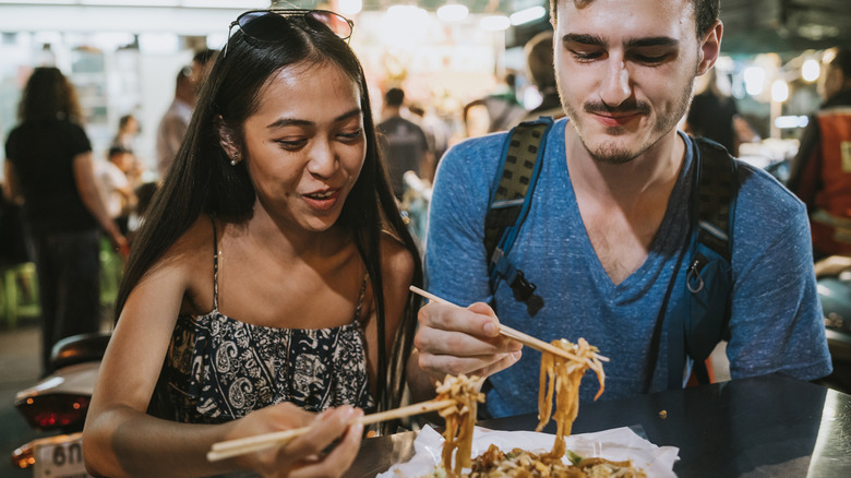 A man and a woman eating drunken noodles with chopsticks at a Thai night market