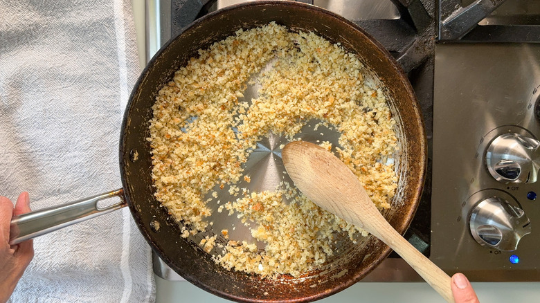 Breadcrumbs being toasted in saute pan with avocado oil and being stirred with wooden spoon