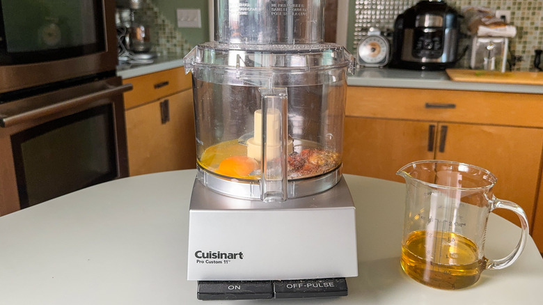 Egg yolks, spices, vinegar, and mustard in food processor bowl with measuring cup of oil