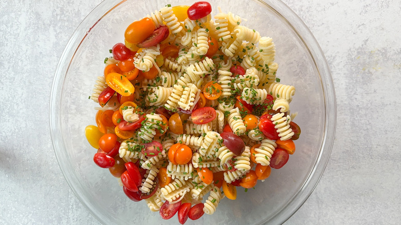 Cooked radiatore pasta, chopped chives, and halved grape and cherry tomatoes in glass bowl on countertop