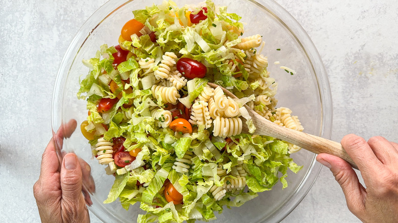 Folding chopped romaine lettuce into cooked radiatore pasta and tomato halves with wooden spoon in glass bowl