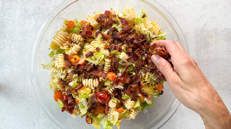 Adding chopped cooked bacon to over-the-top BLT pasta salad in glass bowl