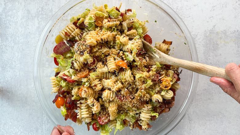 Stirring toasted breadcrumbs into over-the-top BLT pasta salad in glass bowl with wooden spoon