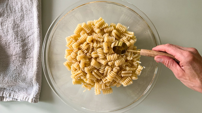 Adding avocado oil with tablespoon measure to radiatore pasta in glass bowl