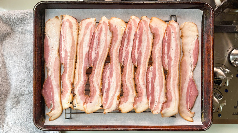 Raw bacon slices on rack over baking sheet pan on stovetop