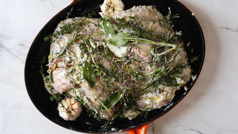 Overhead view of uncooked leg of lamb in a roasting pan with sauce and herbs