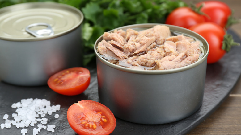 An open can of tuna packed in water with tomatoes, salt, and lettuce in background