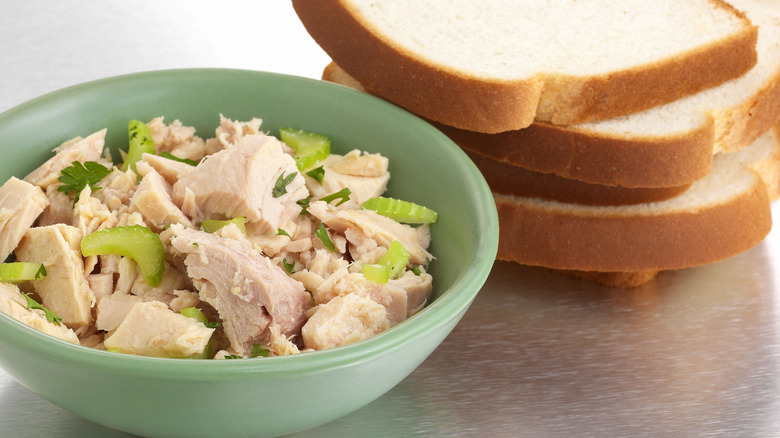 A bowl of tuna salad with big chunks of tuna and celery next to slices of white bread