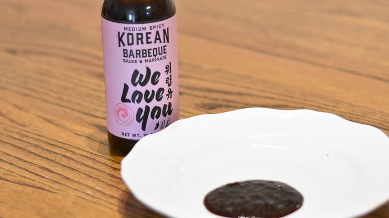 A bottle of medium spicy Korean barbecue sauce from We Love You next to a white plate with a sample on a wooden table