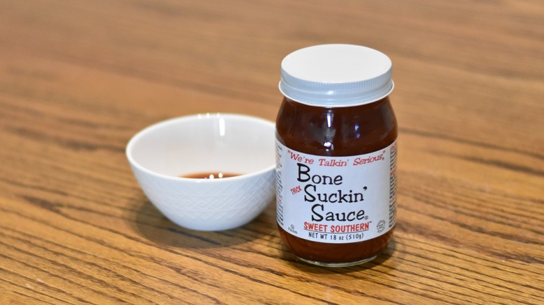 A bottle of Bone Suckin' Sauce next to a white bowl with sample on a wooden table