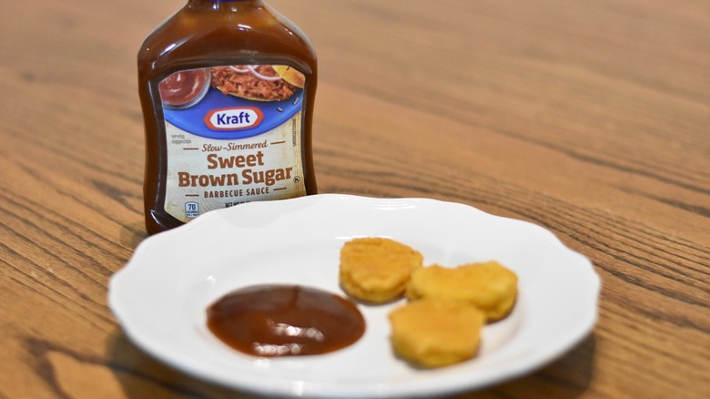 A bottle of Kraft slow-simmered sweet brown sugar barbecue sauce next to a white plate with a sample and three chicken nuggets on a wooden table