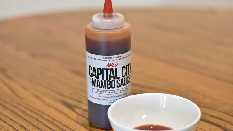 A bottle of Capital City mambo sauce next to a white bowl with a sample on a wooden table