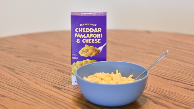 Box of Trader Joe's cheddar macaroni and cheese is next to a blue bowl of macaroni with silver spoon.