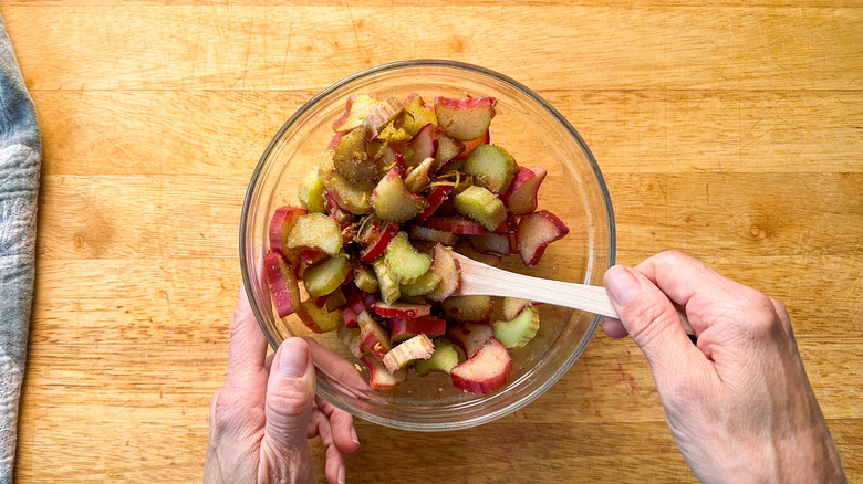 Combining sliced rhubarb with cinnamon, cardamom, and brown sugar in a glass bowl with wooden spoon