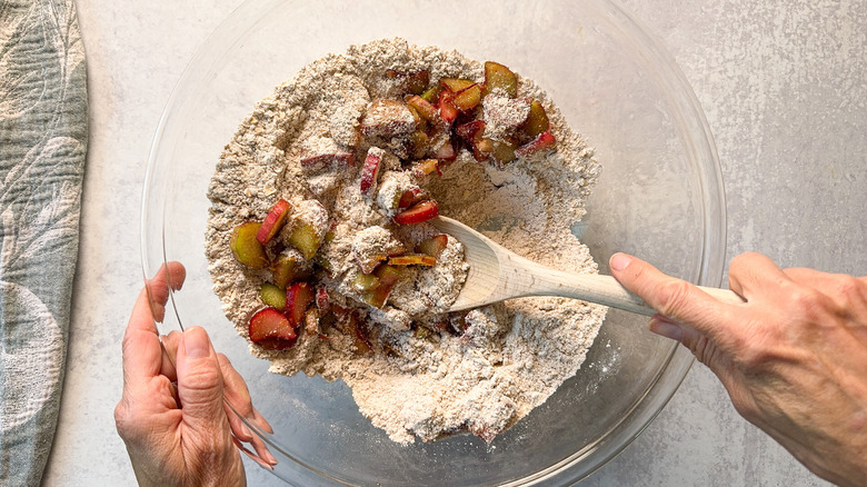 Stirring sliced rhubarb into dry ingredients for rhubarb and cardamom oat muffins in glass bowl with wooden spoon