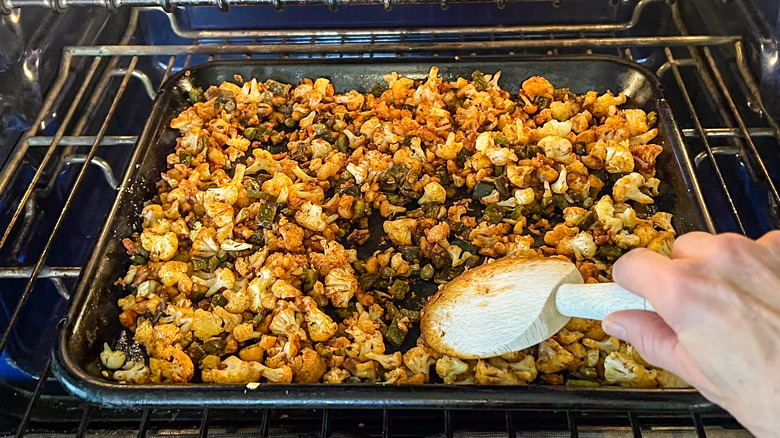 Stirring roasted cauliflower and poblano peppers with spices, walnuts, and capers on baking sheet in oven with wooden spoon