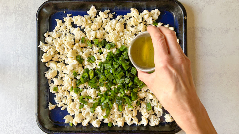 Pouring avocado oil on chopped cauliflower and poblano peppers on baking sheet