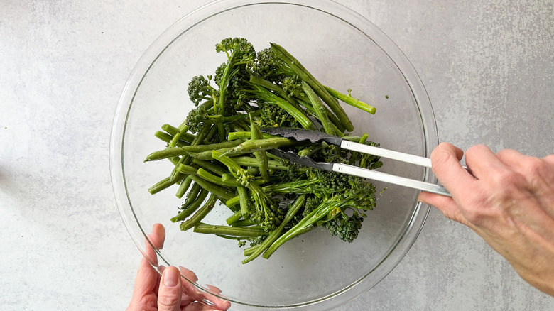 Tossing broccolini and green beans with oil, salt, and pepper in glass bowl with tongs