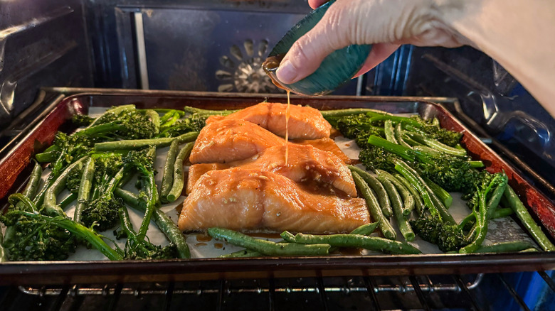 Adding marinade to roasted miso maple salmon with broccolini and green beans on baking sheet in oven