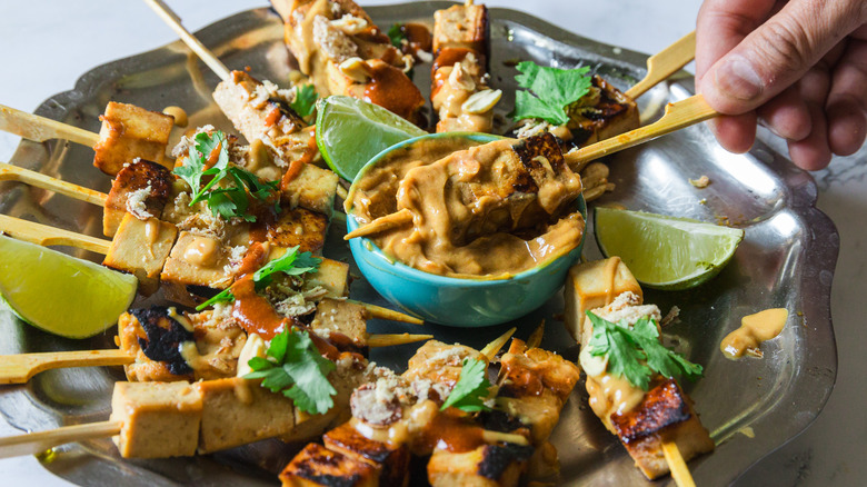 Hand dipping a satay tofu skewer into peanut sauce on a metal plate