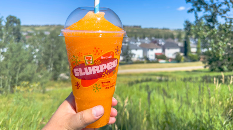 A person holds a cup of 7-Eleven slurpee with orange icy drink inside in front of a grassy field