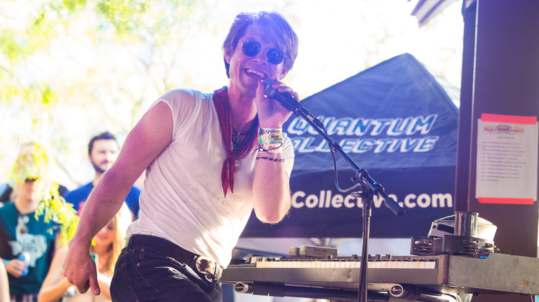 A man wearing a white t-shirt, red bandana, and sunglasses sings into a microphone