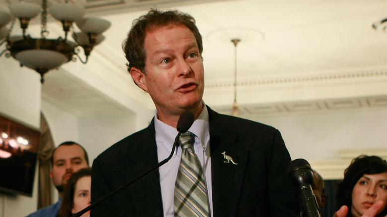 Whole Foods CEO John Mackey speaks at a podium wearing a black suit