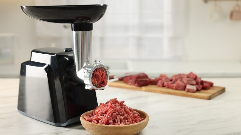 A machine grinds meat into a bowl with more cut-up meat waiting in the background