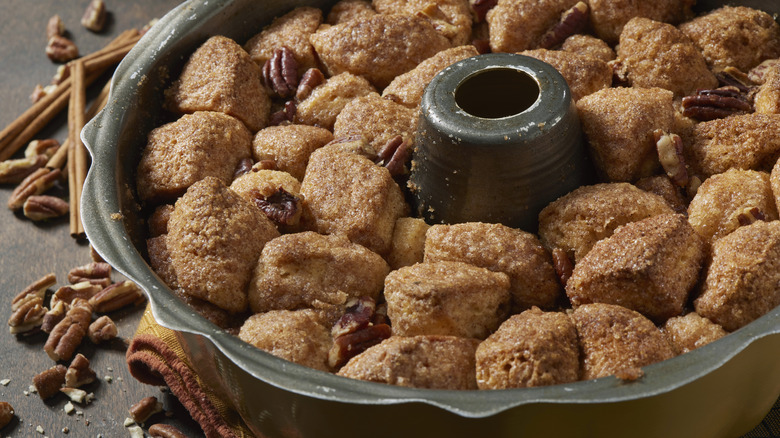 A bundt pan of cinnamon sugar monkey bread topped with pecans, and cinnamon sticks below the pan on the surface of a grey countertop.