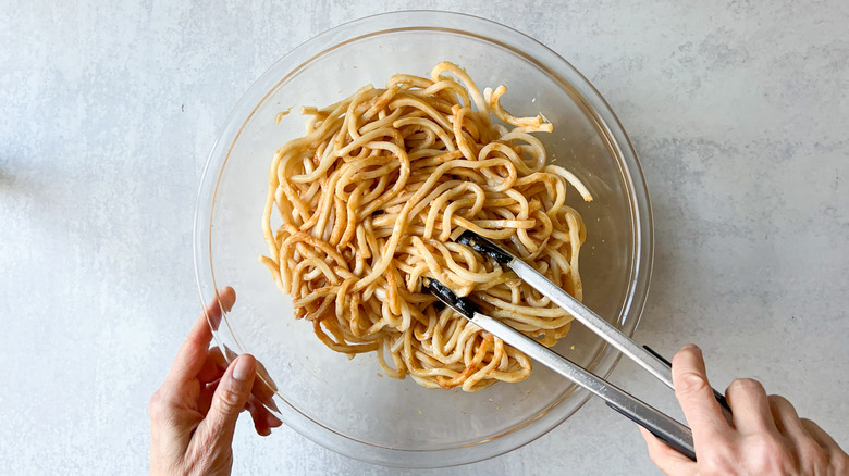 Udon noodles in bowl tossed with dark soy and sesame with tongs