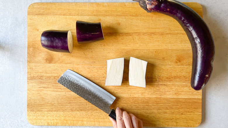 Chinese eggplant sections being halved with knife on cutting board