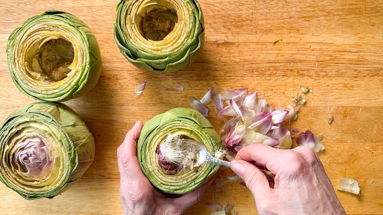 Scooping fuzzy chokes out of fresh artichokes on cutting board with spoon