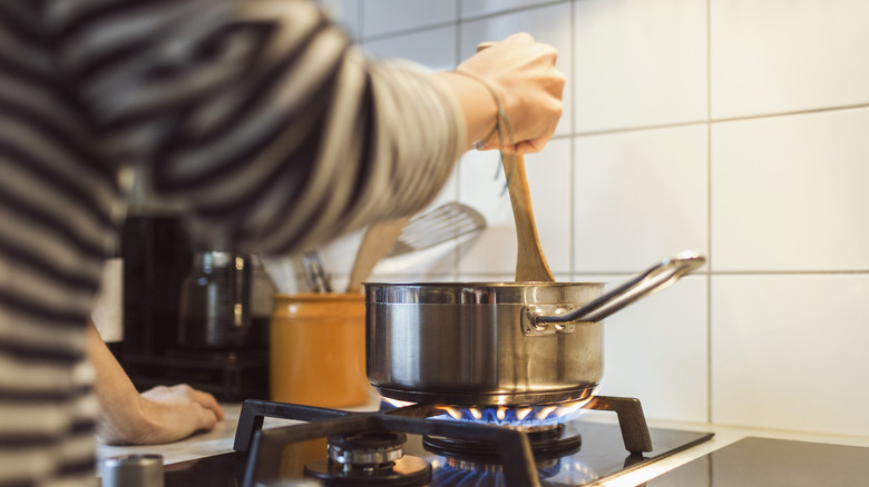 A person stirs a stainless steel saucepan on a gas stove at home.